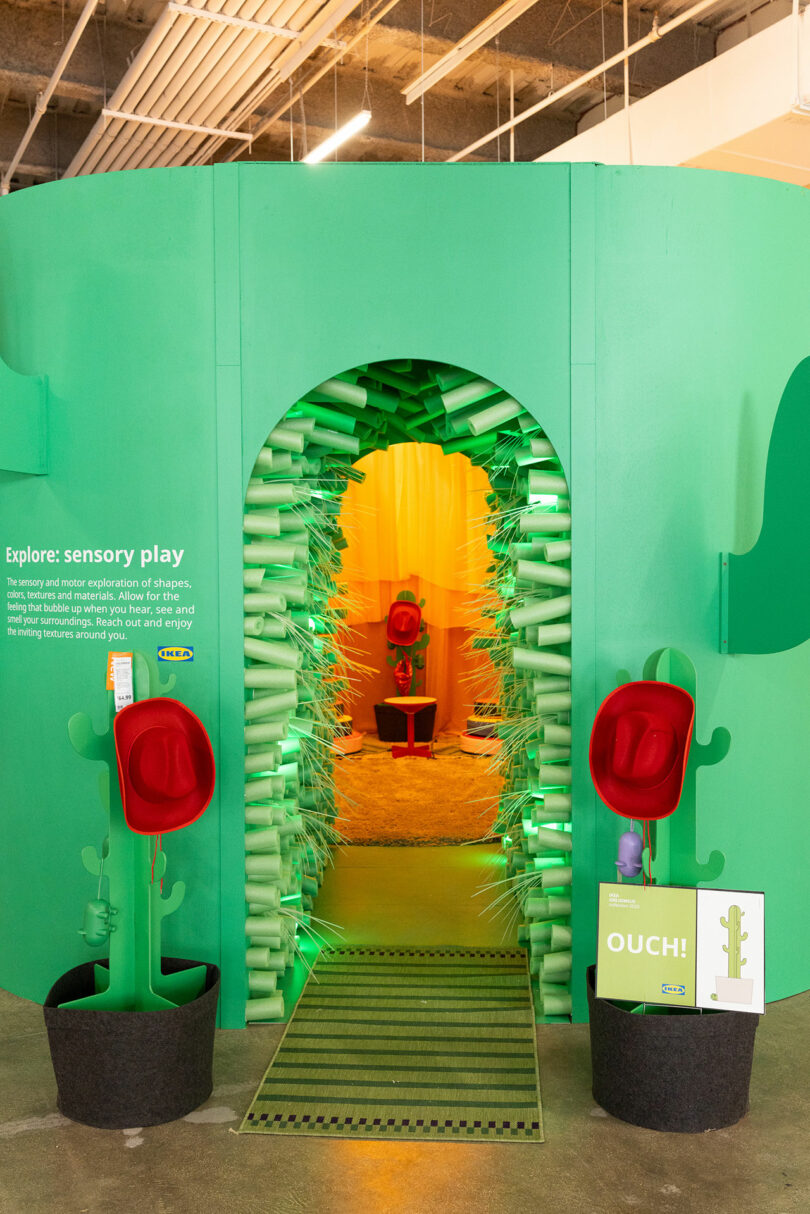 A green sensory play exhibit includes an arch with soft spikes, red hat-like objects, green cactus-shaped decorations, and signage that invites children to explore tactile experiences.