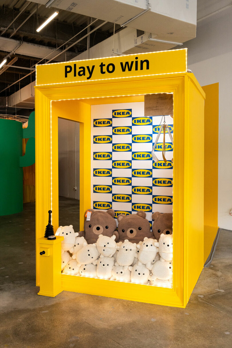 A yellow claw engine room with "Play to win" sign, IKEA logos inside and teddy bear toys stacked up front.