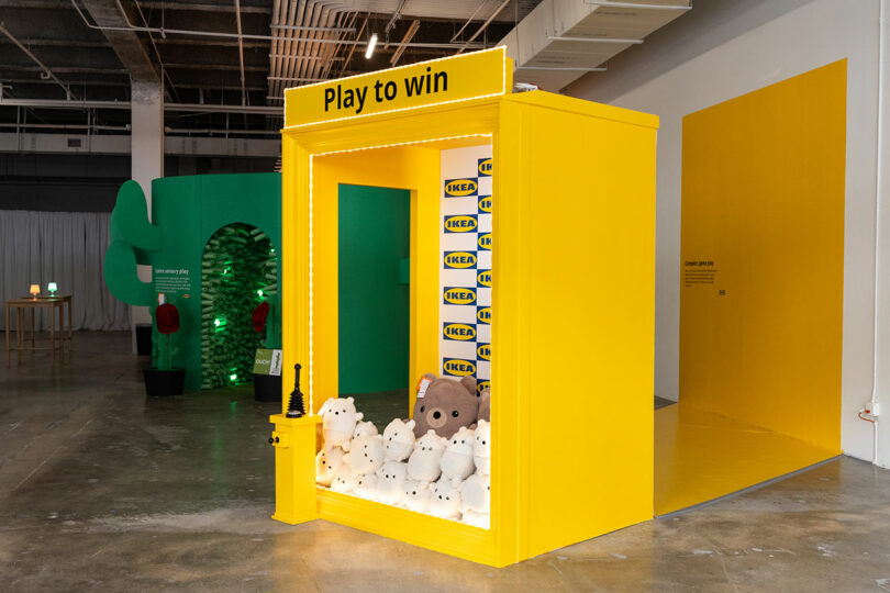 A bright yellow claw machine with a tag "Play to win" filled with IKEA plush toys sits in a spacious interior with industrial ceilings.