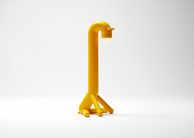 A yellow lamp in the shape of a dog with a rounded body, four short legs and a light source at the end of its curved neck, on a plain white background.