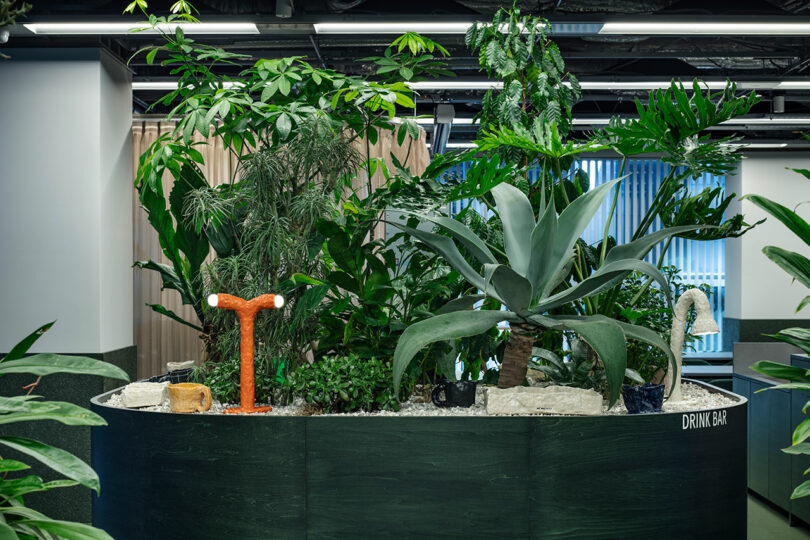 Gran macetero interior lleno de diversas plantas tropicales y decoraciones ubicadas en un moderno espacio de oficina etiquetado "Barra de bebidas.