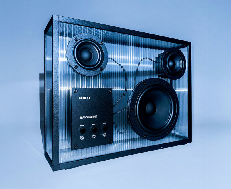 Altavoz transparente con marco negro que muestra sus componentes internos, incluidos tres conos de altavoz y perillas de control, sobre un fondo azul.