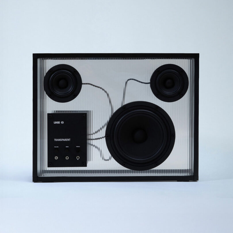 Un altavoz transparente con marco negro y cableado interno claramente visible, tres controladores redondos y un panel de control con la etiqueta 