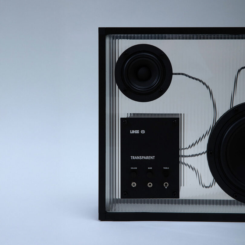 Primer plano de un altavoz transparente con sus cables internos, panel de control y puerto de entrada de audio visibles, sobre un fondo liso.