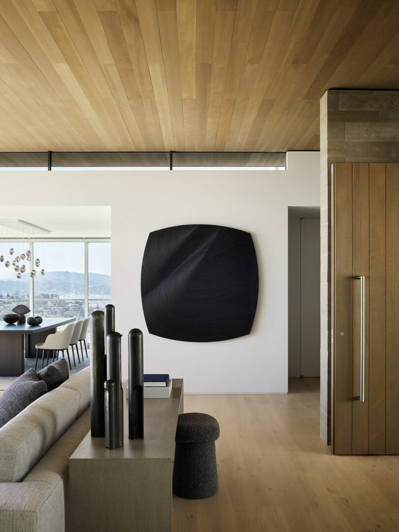 Salón moderno con suelo y techo de madera clara, sofá gris, jarrón decorativo negro sobre la consola y grandes obras de arte abstracto oscuro sobre paredes blancas.