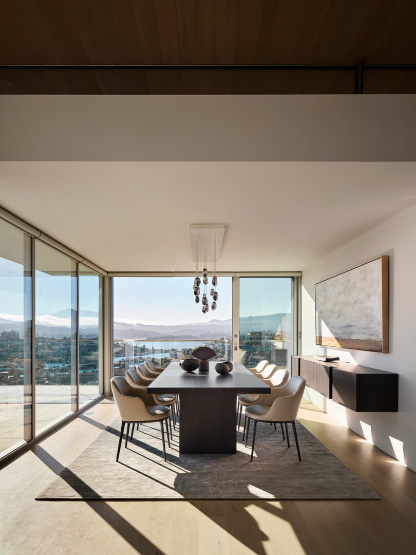 El moderno comedor cuenta con una mesa larga con diez sillas, grandes ventanales con vistas a la ciudad y la montaña, una decoración minimalista y candelabros modernos.