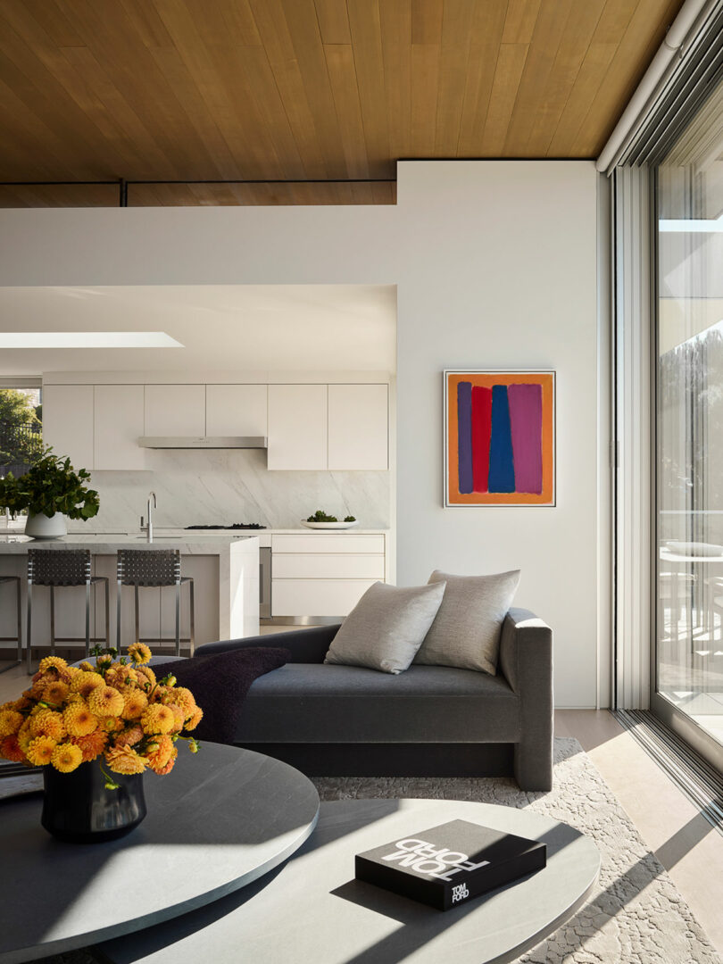 Salón moderno con sillón gris, mesa baja redonda, jarrón naranja, arte abstracto en la pared y vista a la cocina blanca con salpicadero de mármol y taburetes de bar.