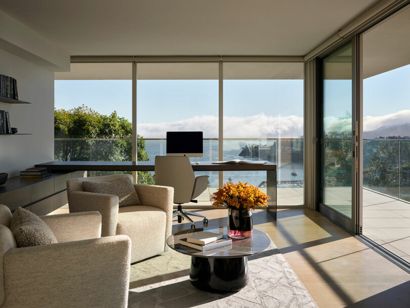 Oficina en casa moderna con grandes ventanales, escritorio, silla de oficina, estanterías, dos sillones, una mesa de café llena de flores y vistas al balcón y al agua exterior.