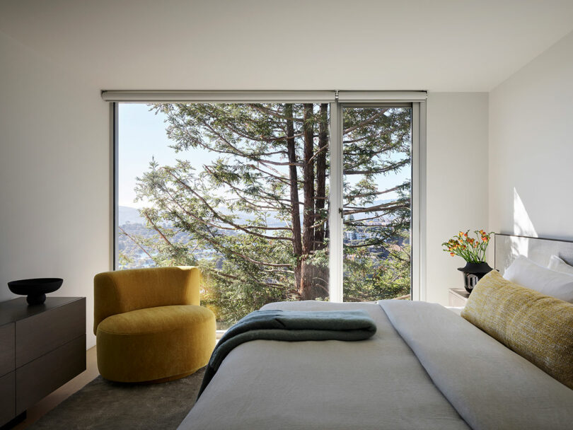 Dormitorio moderno con una ventana grande y un árbol afuera, sillas amarillas, una cómoda gris y una cama ordenada con ropa de cama neutra y detalles en amarillo.