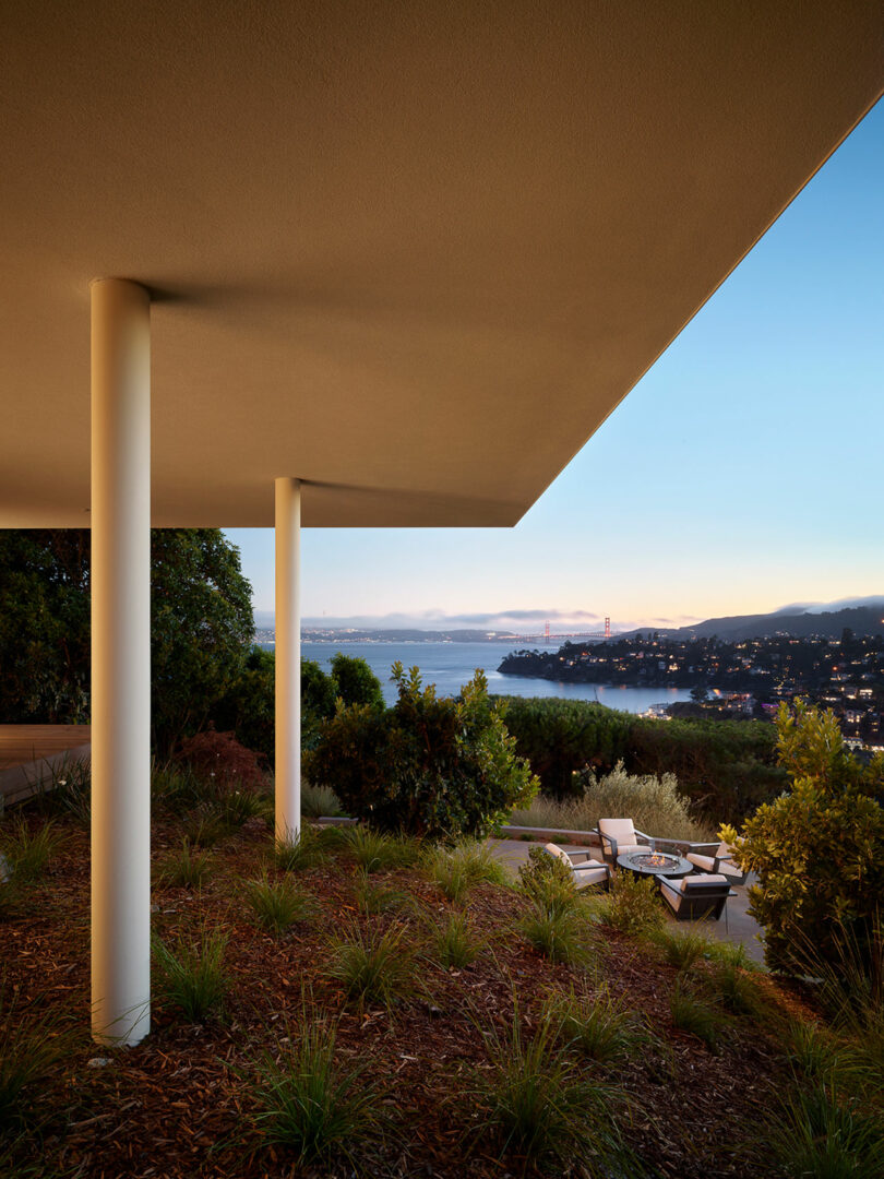 El moderno patio cuenta con columnas blancas y un techo sobresaliente, con vistas a los jardines, el agua y el paisaje urbano lejano al atardecer.