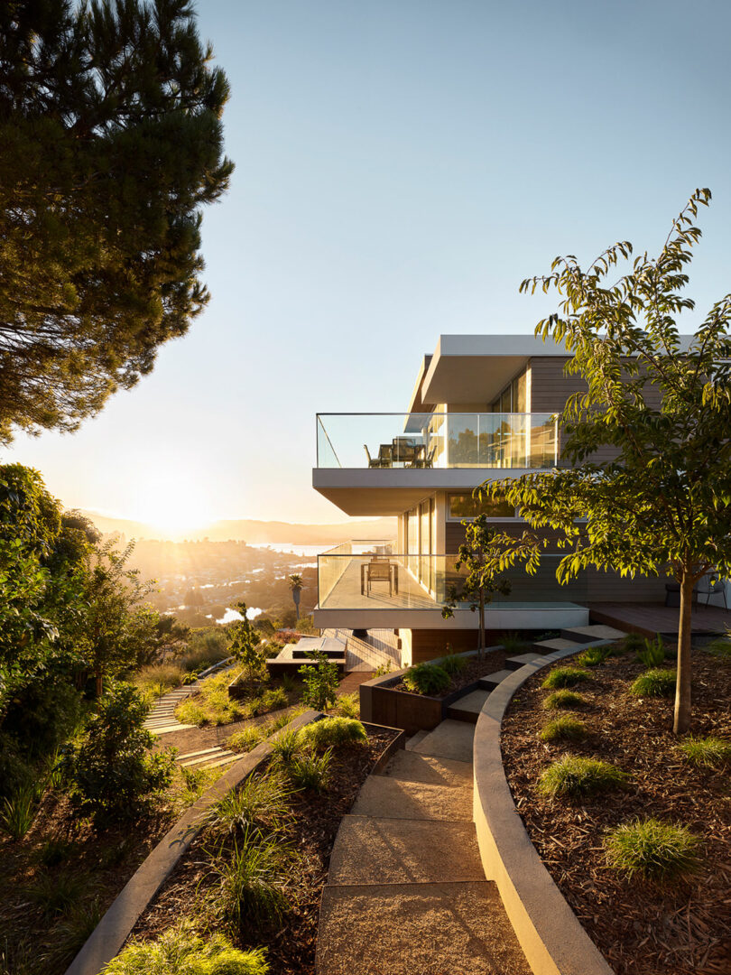 La casa moderna cuenta con un gran balcón de cristal con vistas a la pintoresca ladera al atardecer, con senderos pintorescos y vegetación en primer plano.