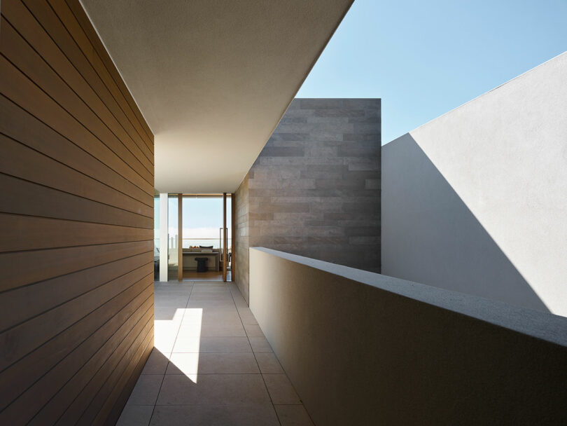 Pasillo moderno y minimalista con paredes de madera y hormigón, suelos de baldosas, luz del sol que proyecta sombras geométricas y conduce a puertas de cristal con vistas al exterior.