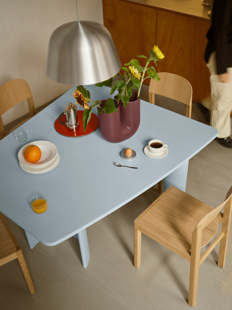 Sobre la mesa azul claro hay un jarrón con girasoles, una taza de café, un huevo, frutas, un vaso de jugo de naranja y una cafetera de metal debajo de la lámpara de araña plateada.