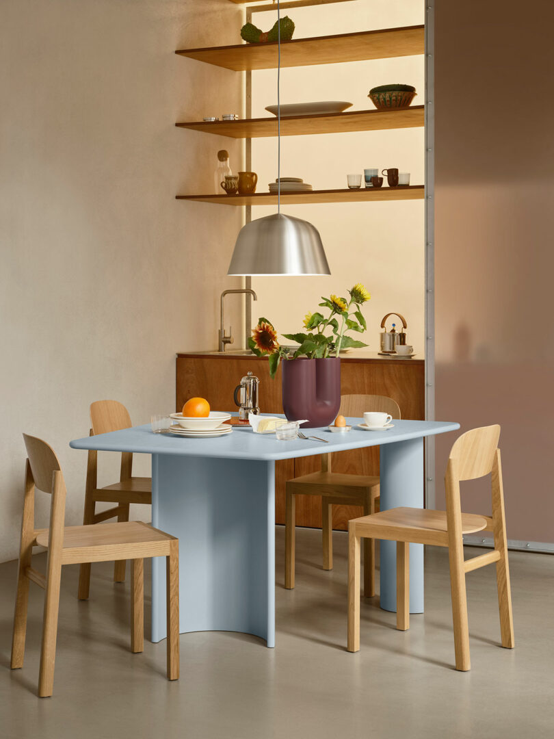 Comedor moderno con una mesa azul claro, cuatro sillas de madera, un jarrón con girasoles, artículos para el desayuno y estantes abiertos con platos al fondo.