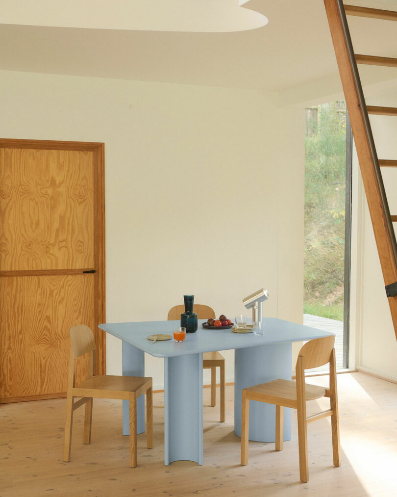 Un comedor sencillo, una mesa azul claro, tres sillas de madera, vajilla variada y frutas, una puerta de madera, una escalera y grandes ventanales con árboles verdes en el exterior.