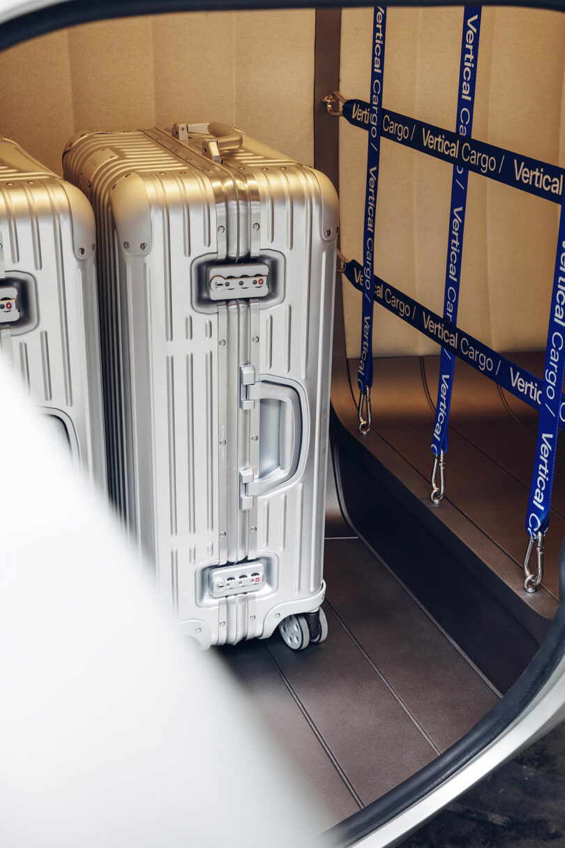 Dos maletas rígidas plateadas se colocan en el maletero de un automóvil junto a correas de carga verticales etiquetadas "carga vertical.