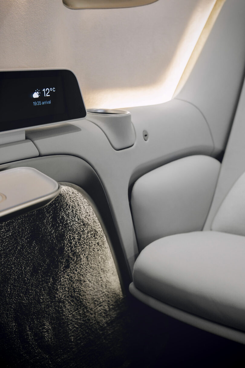 Primer plano del asiento de un avión moderno en una cabina premium con pantalla digital que muestra 12 °C y la hora de llegada, con un interior elegante y luminoso.