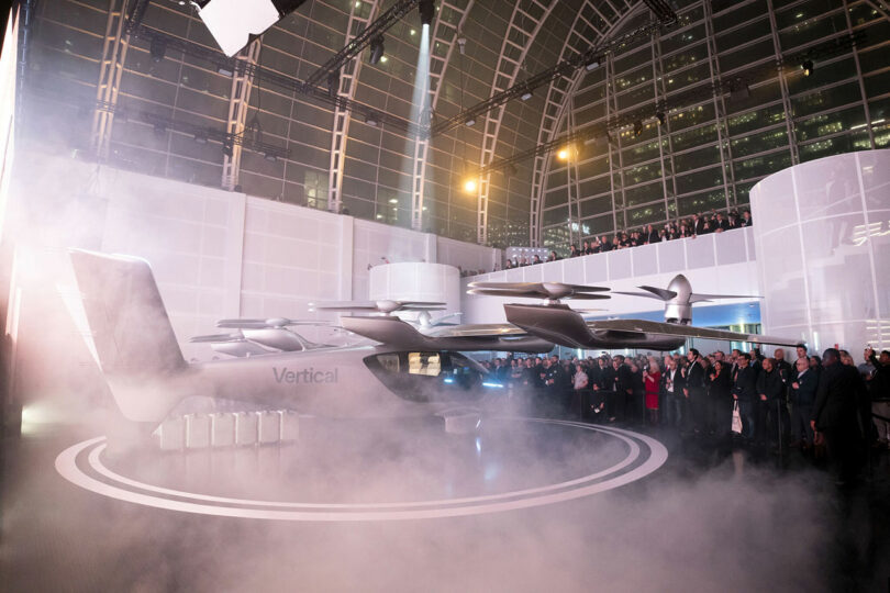 Una gran multitud de personas observa un avión de despegue y aterrizaje vertical (VTOL) en exhibición en el interior, rodeado de niebla y arquitectura moderna.