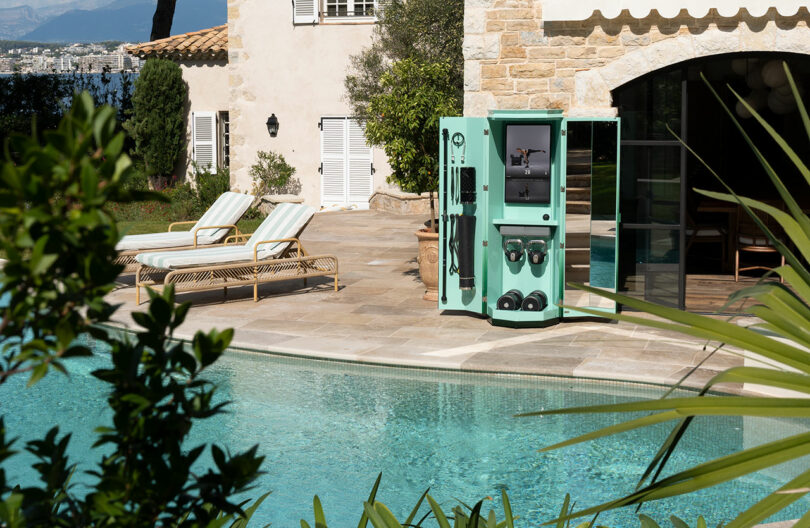 A home gym cabinet with workout equipment stands open beside a swimming pool on a stone patio outside a house.