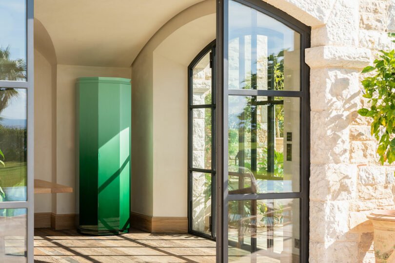 A modern green refrigerator stands in a sunlit corner of a stone house with large glass doors and windows.