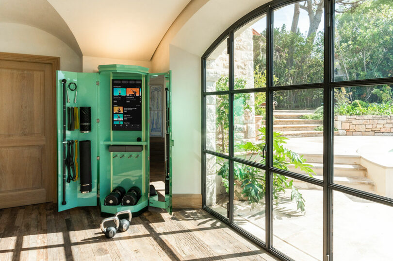 A compact home gym cabinet with exercise equipment and a digital workout screen sits near a large arched window overlooking a garden.