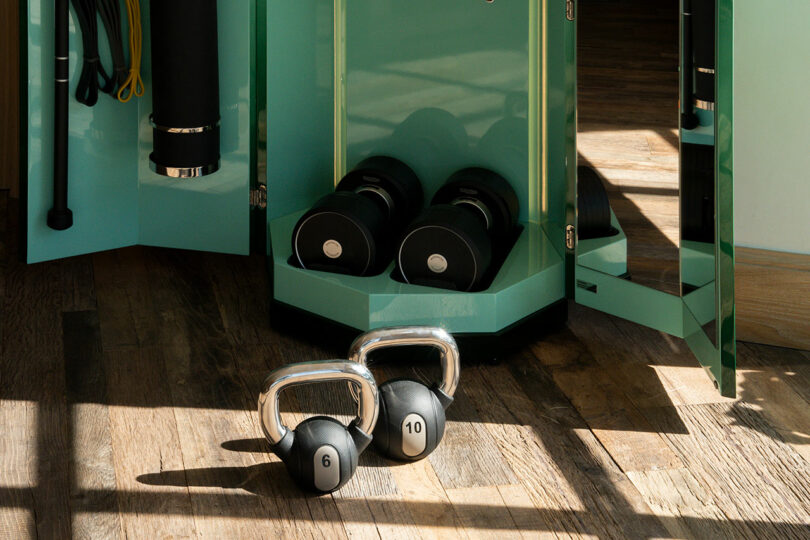 Two kettlebells labeled 6 and 10 sit on a wooden floor in front of a cabinet containing two dumbbells and exercise equipment.