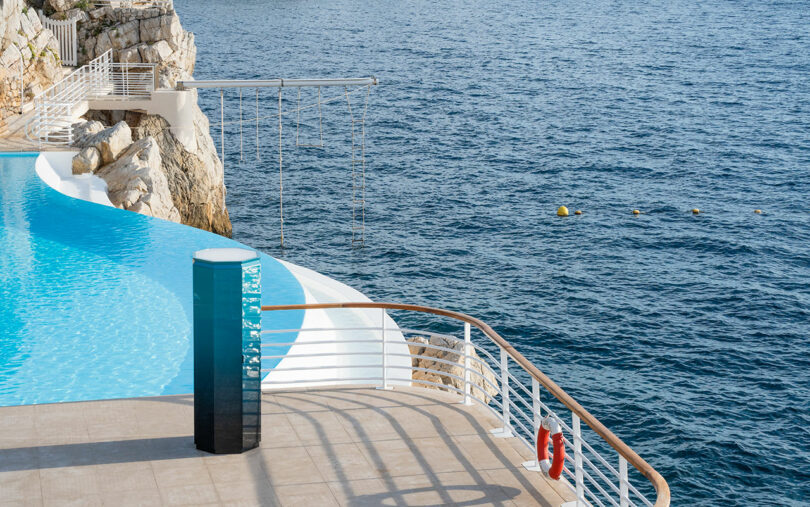 Outdoor infinity pool overlooking the sea, with safety railing, lifebuoys, steps leading to the water, and rocky cliffs in the background.