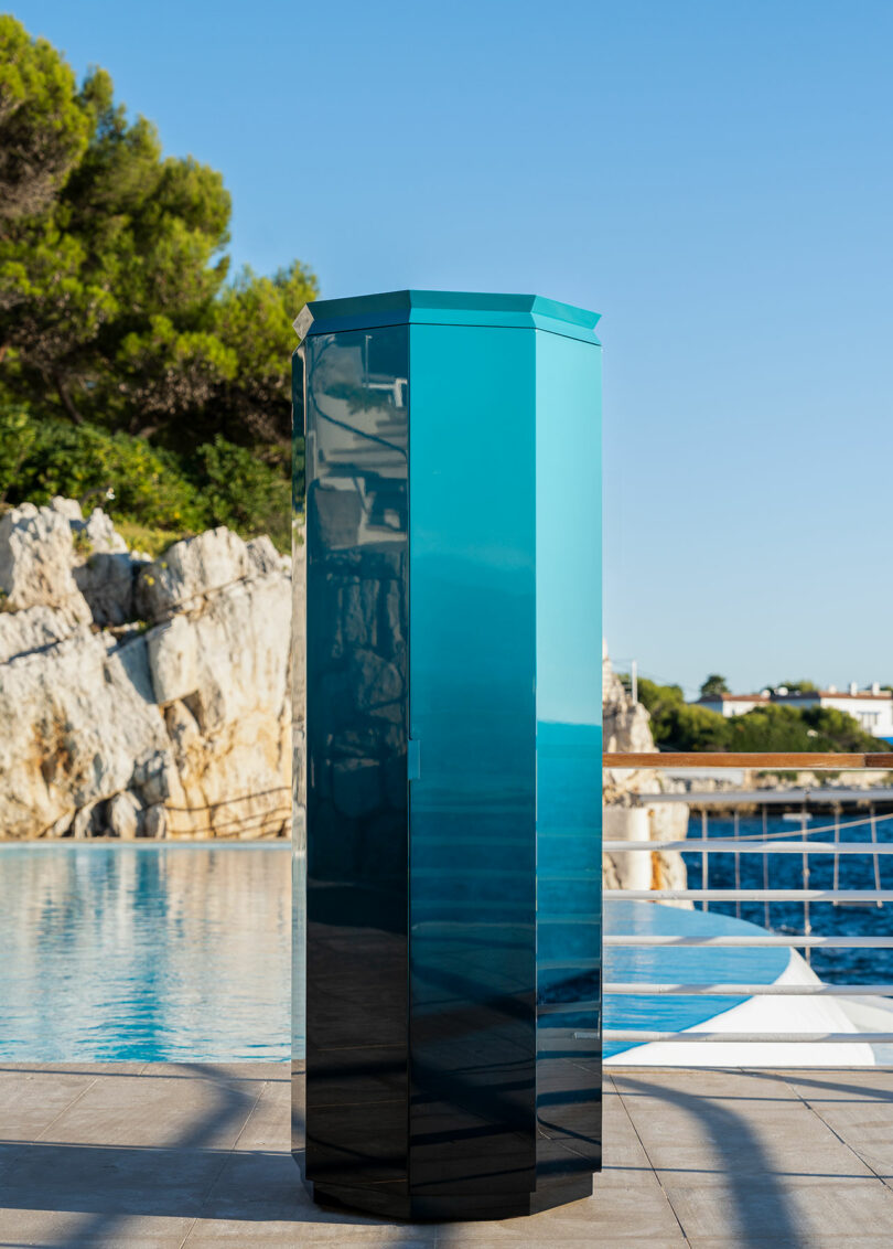 A tall, glossy, blue-green geometric sculpture stands on a tiled patio near a pool, with rocky cliffs and trees in the background.