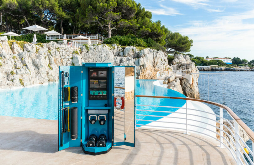 A fitness equipment locker stands open on a poolside terrace overlooking the sea, with rocky cliffs, trees, and lounge chairs in the background.