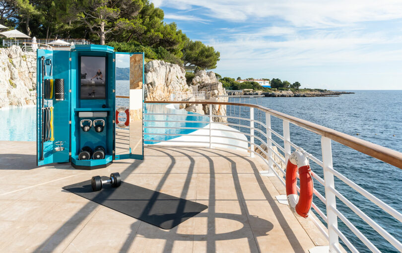 A compact outdoor gym setup with equipment and a mat is positioned on a terrace overlooking the sea, with railings, safety rings, and coastal scenery in the background.