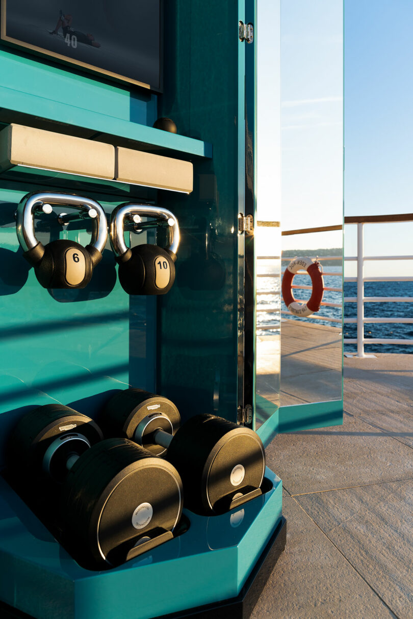 A compact gym setup on a yacht deck features kettlebells, adjustable dumbbells, and a mirror with an ocean view in the background.