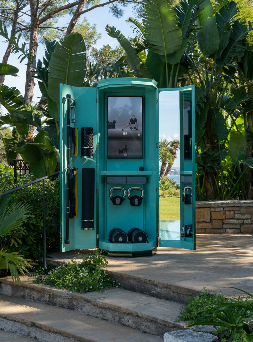 A turquoise outdoor fitness cabinet with open doors reveals exercise equipment, weights, and a built-in screen, surrounded by tropical plants on a stone patio.