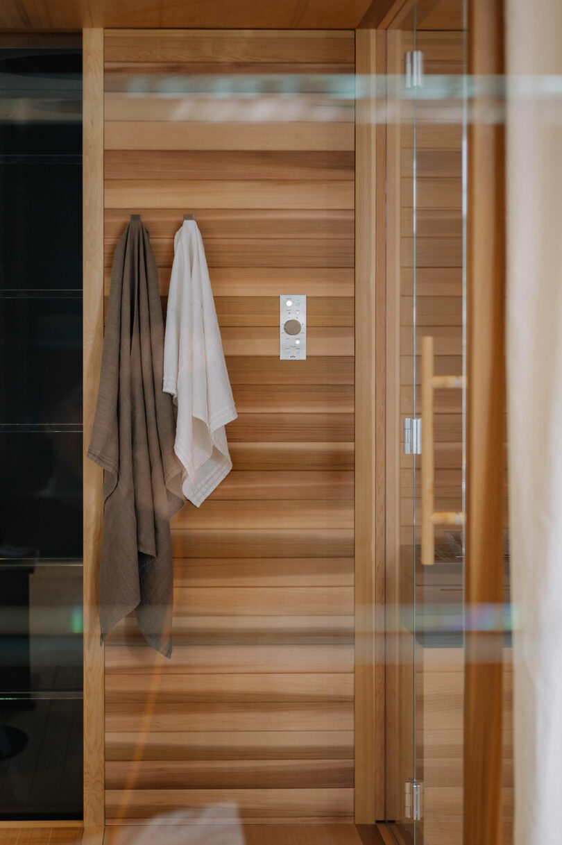 Wood-paneled sauna interior with a white and a gray towel hanging on hooks next to a control panel and a glass door.