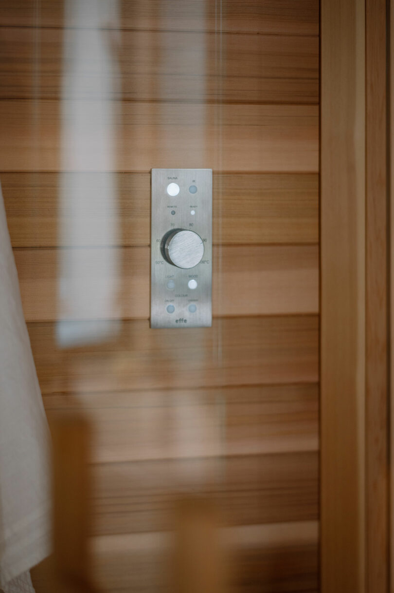 A close-up of a silver control panel with buttons and a knob mounted on a wooden wall, likely inside a sauna or wooden room.