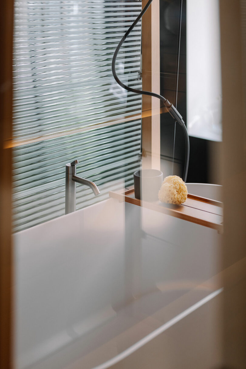 A modern bathtub with a wooden tray holding a yellow sponge and a cup, set next to a silver faucet and a handheld showerhead.