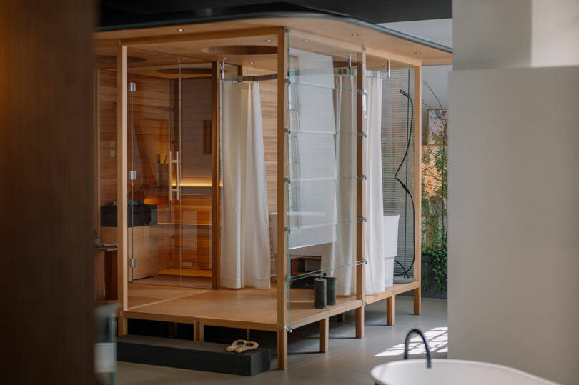 Modern indoor sauna and shower area with wooden and glass construction, white curtains, and a freestanding bathtub nearby in a minimalist bathroom setting.