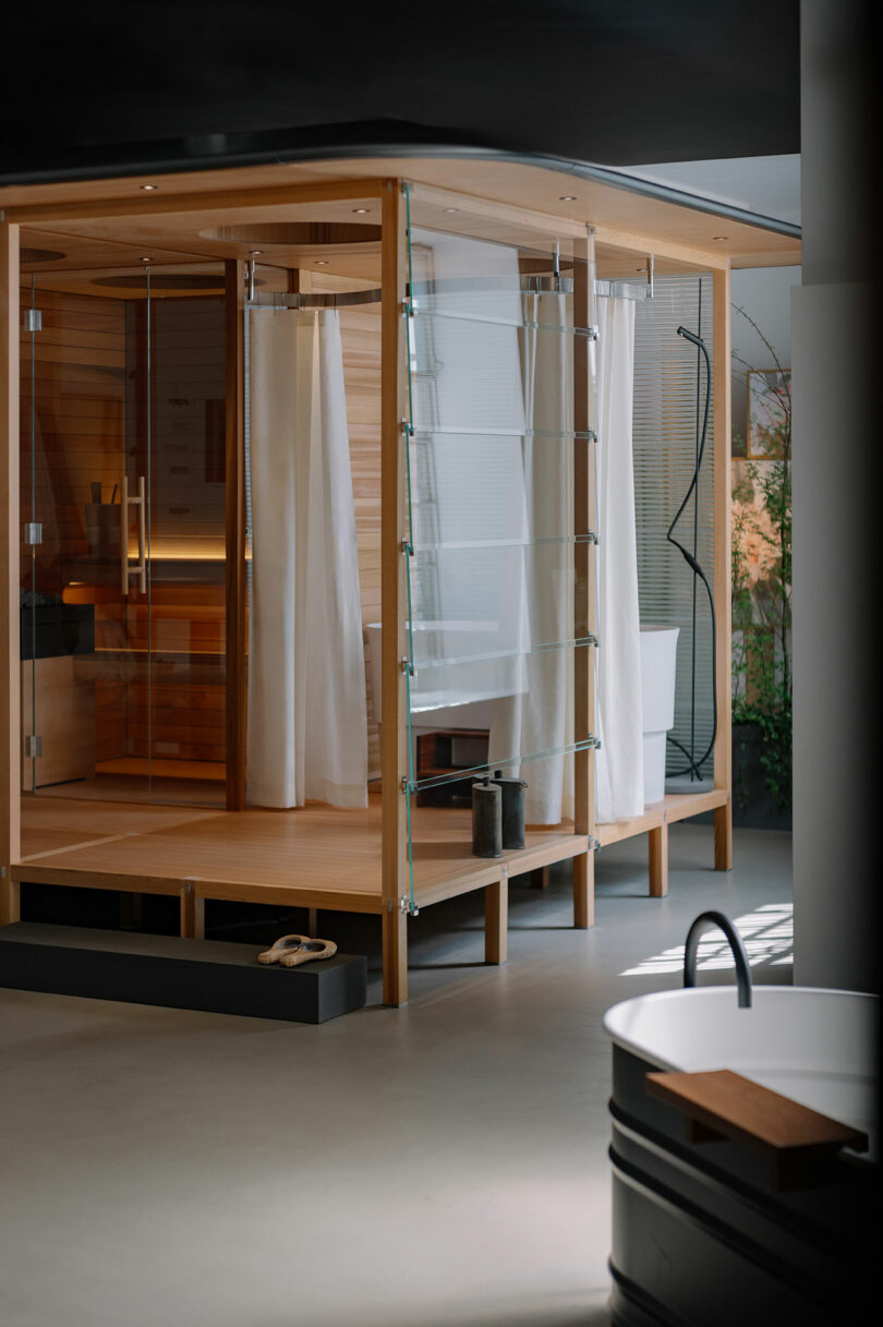 Modern bathroom with a wooden sauna, glass walls, white curtains, black shower area, and a freestanding black bathtub in the foreground.