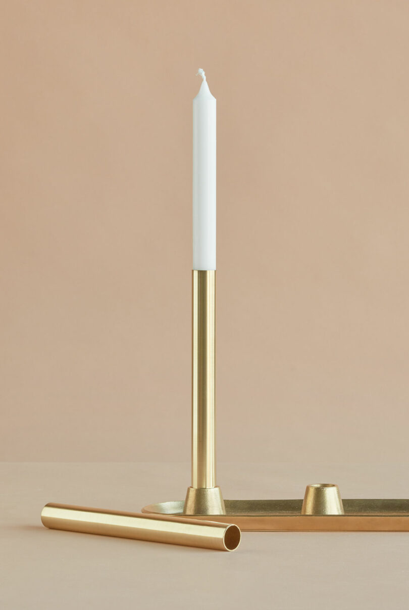 A white candle in a gold holder stands upright on a gold tray, with a separate gold tube lying in front of it on a beige surface.