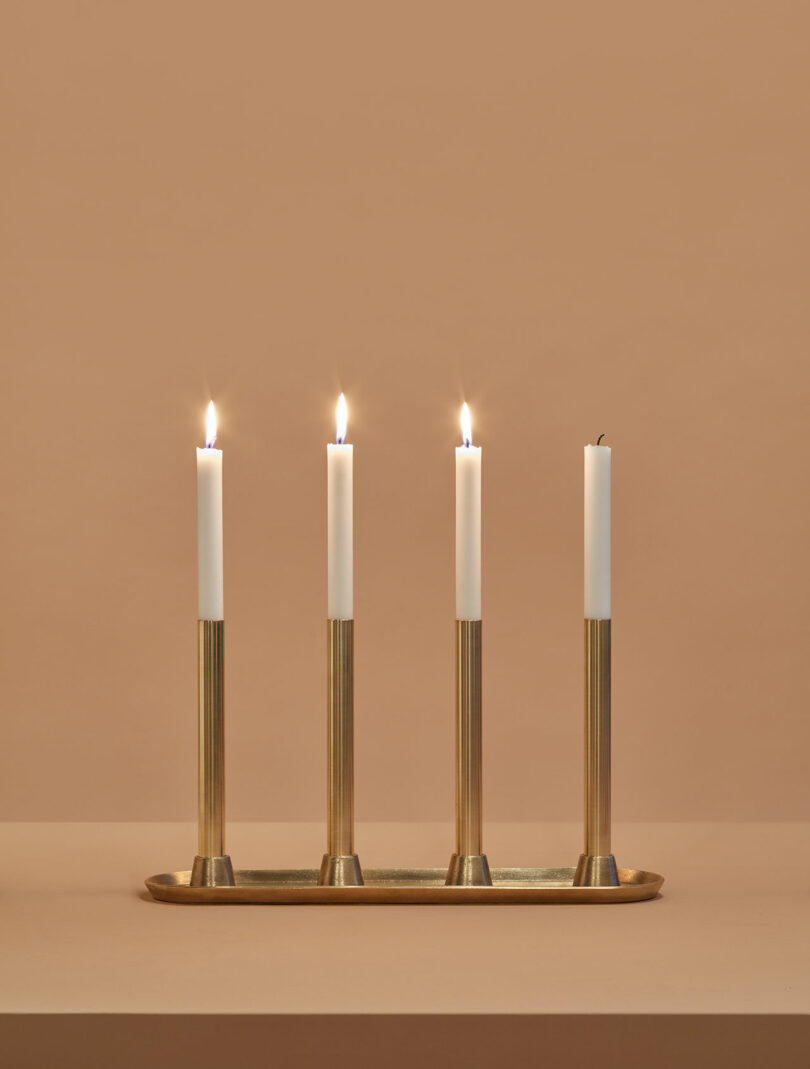 Four brass candle holders with white candles on a brass tray; three candles are lit while the fourth is unlit, against a plain beige background.