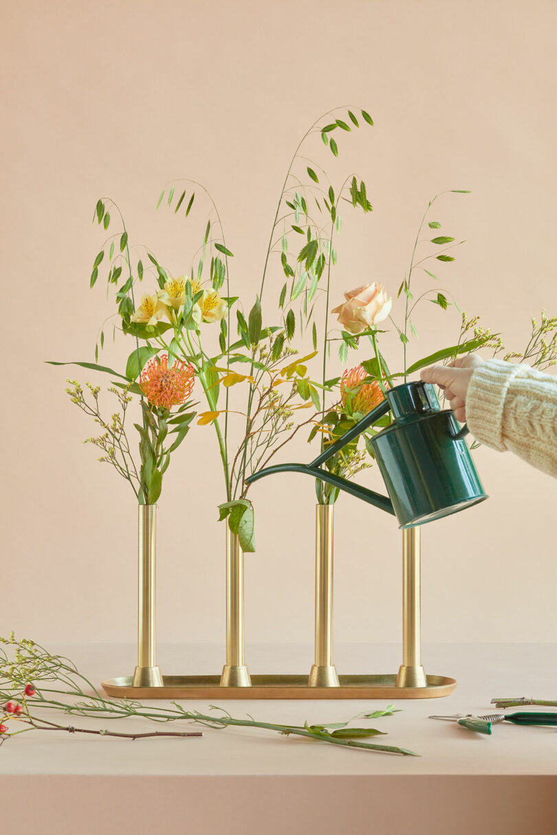 A hand pours water from a green watering can into a modern gold vase holding assorted fresh flowers and greenery on a peach background.