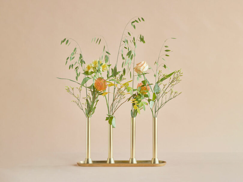 Four brass vases on a matching tray, each holding a minimal arrangement of green foliage and a few orange and yellow flowers, set against a plain beige background.