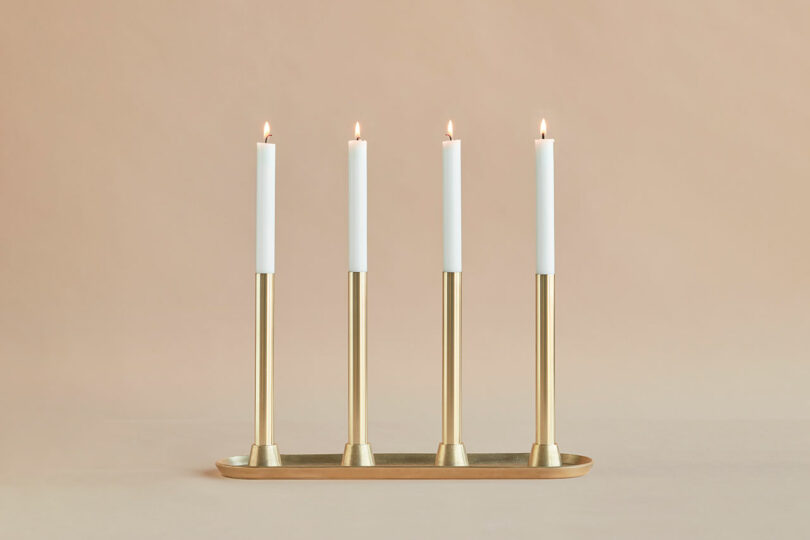 Four lit white candles in gold candleholders are arranged in a row on a matching gold tray against a neutral background.