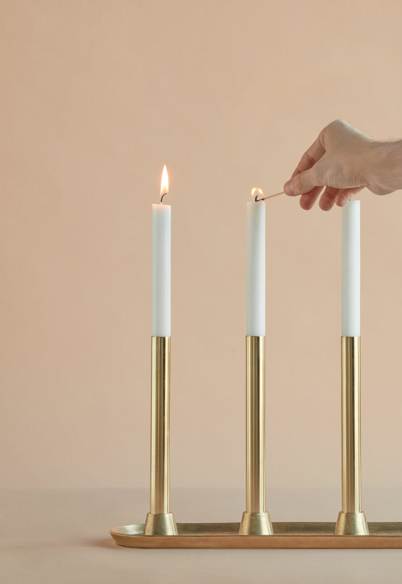 A hand lights the middle of three white candles in gold holders, using a match. The left candle is already lit, against a beige background.