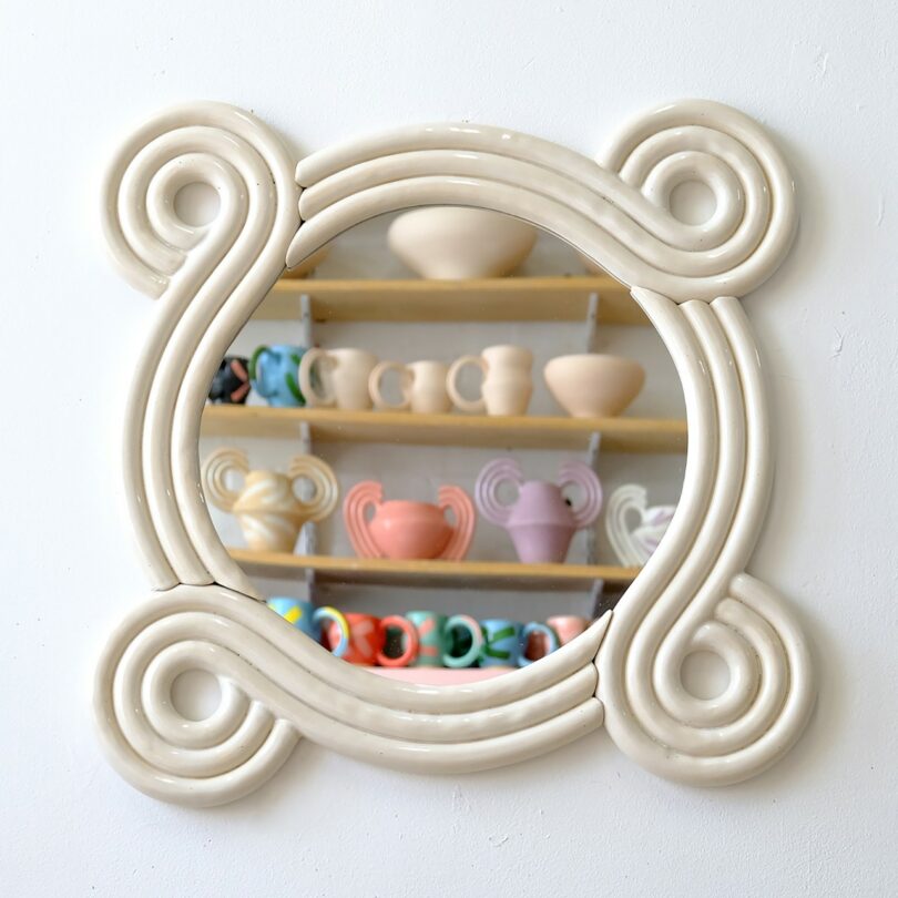 Un espejo redondo con un marco blanco ondulado tallado refleja un estante con coloridas tazas y jarrones de cerámica en una pared blanca