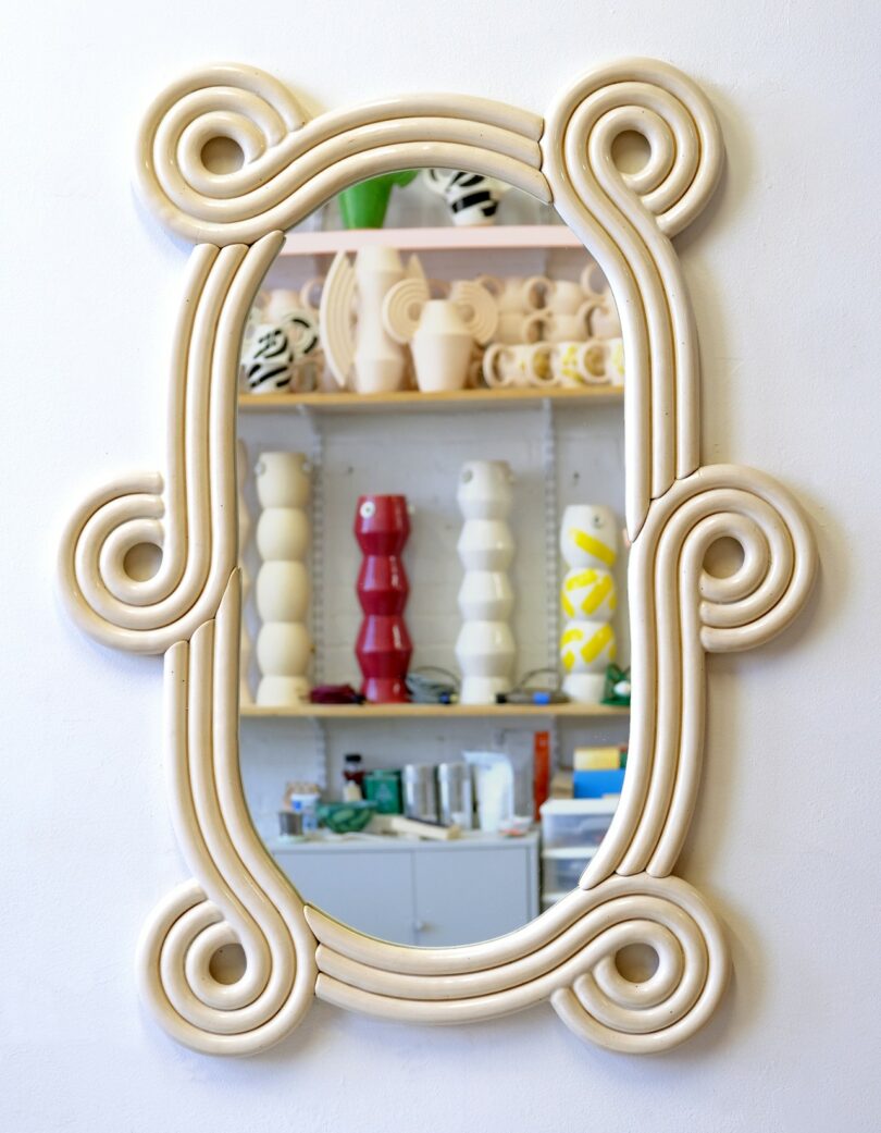 Un gran espejo beige adornado con un patrón de volutas refleja un estante lleno de jarrones esculturales y cerámicas en un ambiente de estudio.