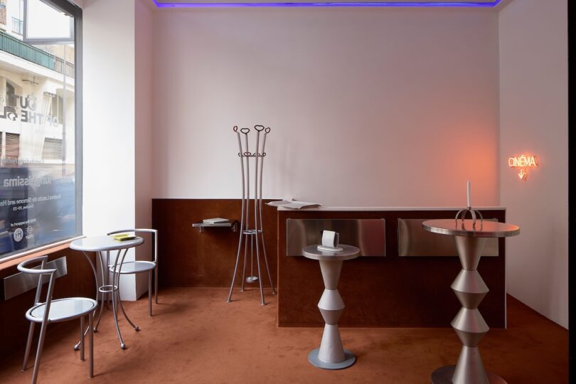 Habitación moderna con muebles geométricos de metal, mesa redonda con dos sillas, mostrador, escultura abstracta, perchero y luces de neón. "Película" Firme sobre la alfombra marrón.