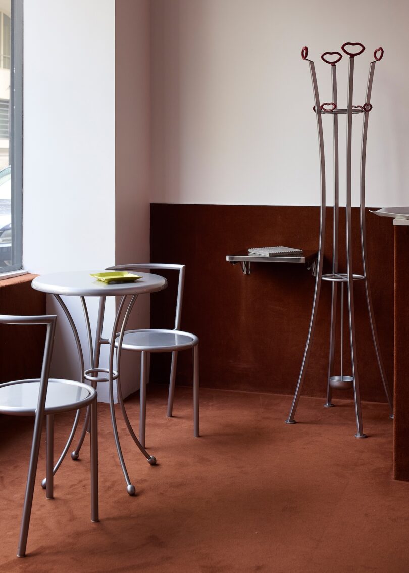 La habitación con alfombra marrón y paredes blancas contiene una pequeña mesa redonda, dos sillas, un escritorio plegable montado en la pared y un perchero alto.