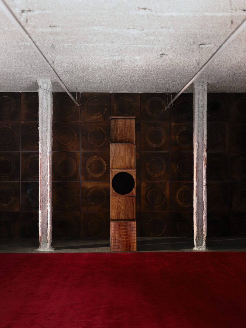 Un alto altavoz de madera se encuentra en el centro de una pared de textura oscura con un patrón circular. En primer plano hay columnas blancas y una alfombra roja.