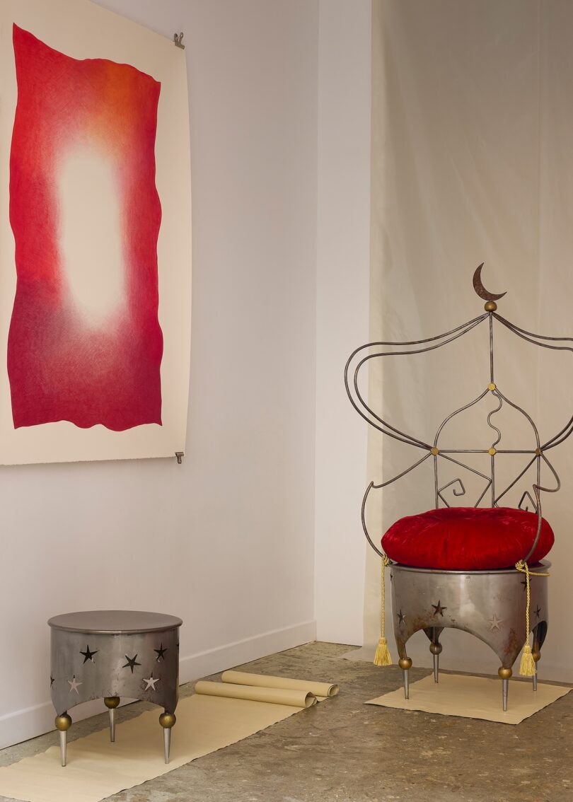 Una silla de metal con cojines rojos y un diseño de luna creciente se encuentra junto a una mesa auxiliar de metal en una habitación minimalista, donde obras de arte abstractas en rojo y blanco cuelgan de la pared.