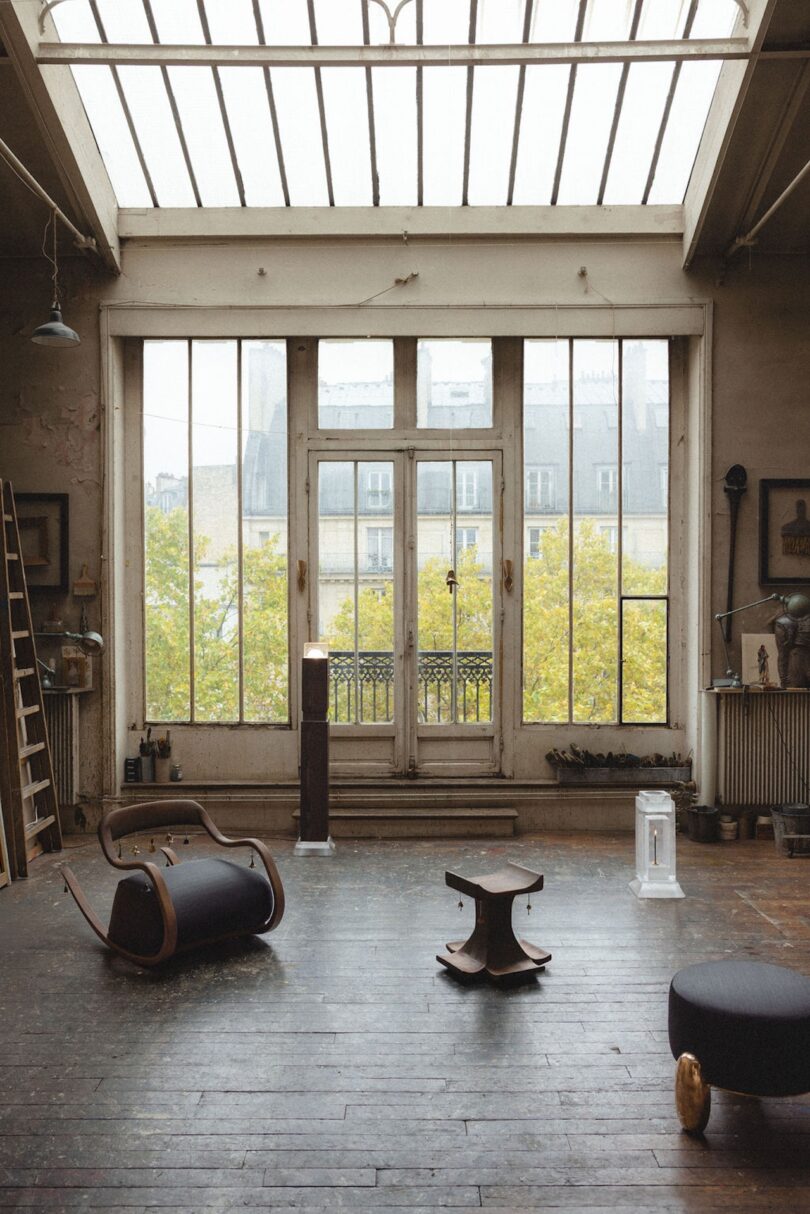 Habitación espaciosa y rústica con un gran tragaluz y ventanas altas, suelos de madera, muebles modernos dispersos y vistas a los árboles y edificios del exterior.